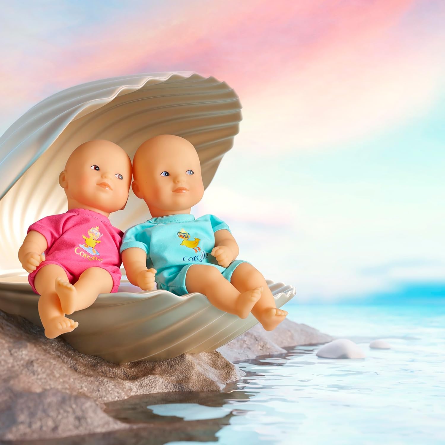 The Corolle Mini Bath Doll - Blue, crafted from soft-touch vinyl, sits in a large open seashell by the water under a pastel-colored sky.