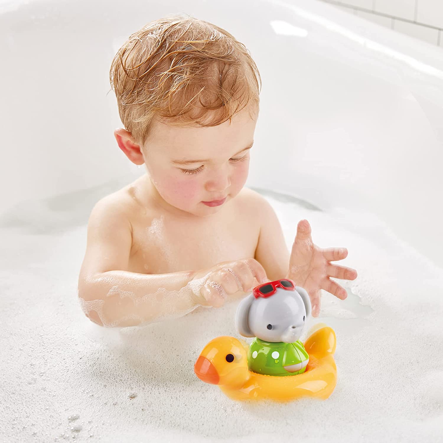 A baby is having bath time, playing with a Spin Splash & Swim Elephant Color-Changing Bath Toy duck in a tub by Hape.