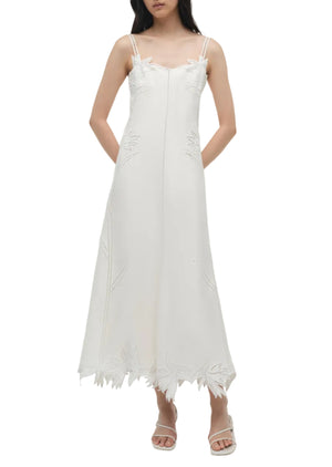 A woman wearing the Simkhai Ellery Bustier Midi Dress—sleeveless with thin straps, subtle leaf patterns, and an uneven hem—paired with white sandals.