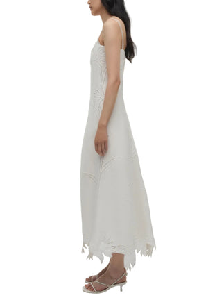 A woman in the Simkhai Ellery Bustier Midi Dress—sleeveless with a floral pattern and scalloped hem—pairs it with strappy sandals, standing in profile against a plain background.