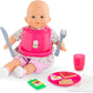 A Corolle baby doll with a pink bib sits with the Large Mealtime Set—featuring accessories like a toy fork, knife, Meal Box plate, packet, pink cup, and napkin.