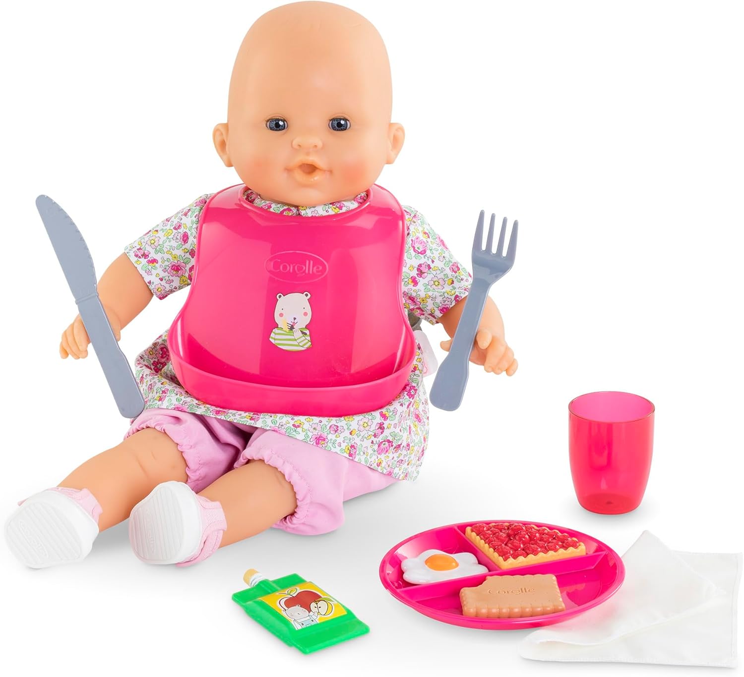 A Corolle baby doll with a pink bib sits with the Large Mealtime Set—featuring accessories like a toy fork, knife, Meal Box plate, packet, pink cup, and napkin.