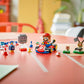 The LEGO® Super Mario™ Mario Kart™ – Interactive LEGO® Mario™ & Standard Kart by Legos - Toyhouse is displayed on a red table, showcasing Mario in his kart, racing features, and checkered flags in a bright room.