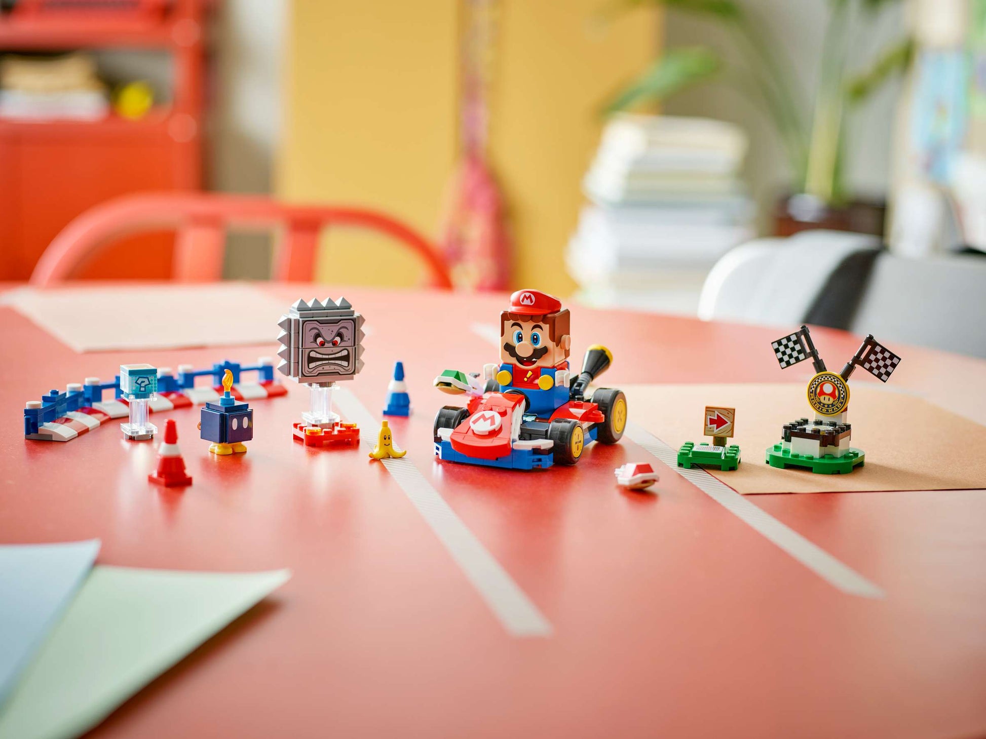 The LEGO® Super Mario™ Mario Kart™ – Interactive LEGO® Mario™ & Standard Kart by Legos - Toyhouse is displayed on a red table, showcasing Mario in his kart, racing features, and checkered flags in a bright room.