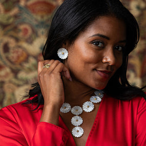 A woman in a red blouse wears Juliska Capucine De Wulf's Mermaid Garden Blossom Stud Earrings and a matching white floral necklace, gently touching her earring as she poses against a patterned background.
