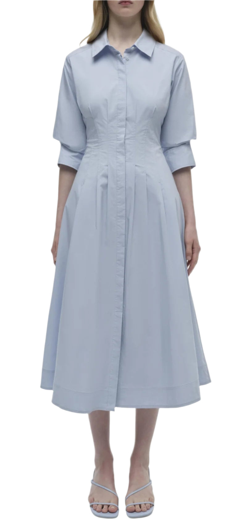 A woman exudes effortless elegance in the Simkhai Signature Jazz Dress, a light blue cotton poplin ensemble featuring a collared design, A-line silhouette with a fitted waist, and mid-length skirt. She completes the look with graceful sandals.