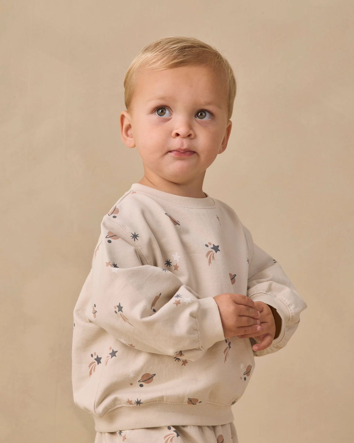 A toddler with short blond hair wears the Quincy Mae Baby Boys’ and Boys’ Oversized Crew Sweatshirt, a cozy fleece pullover adorned with small star and planet prints, standing against a plain beige background—the perfect layering piece for cool days.