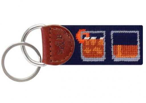 The Smathers & Branson Bourbon Five Ways Key Fob features a charming needlepoint design with cross-stitched images of two glasses: one filled with liquid and an orange garnish, and the other plain. Enhanced with brown leather and embossed initials, this key fob is ideal for adding a splash of color and personality to your pocket or purse.