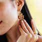 A close-up of a woman wearing Juliska Capucine De Wulf Pathway Post Drop Earrings in a bold geometric style, along with a wide gold ring, as she gently touches her earlobe.