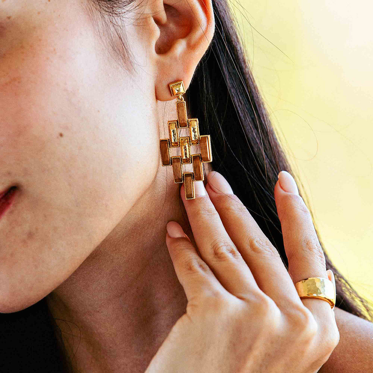 A close-up of a woman wearing Juliska Capucine De Wulf Pathway Post Drop Earrings in a bold geometric style, along with a wide gold ring, as she gently touches her earlobe.
