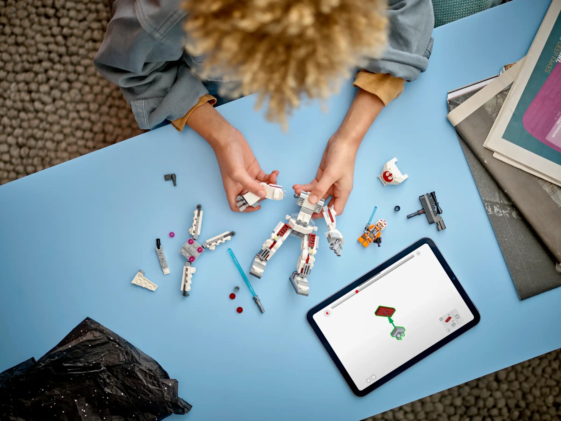 Someone builds the LEGO® Star Wars™ Luke Skywalker™ X-Wing™ Mech from Legos - Toyhouse on a blue table, using various parts, while following instructions displayed on a nearby tablet.