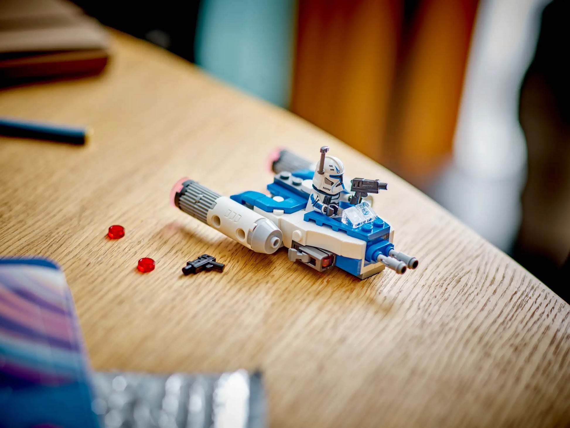A LEGO® Star Wars™ Captain Rex™ Y-Wing™ Microfighter by Legos - Toyhouse features a Captain Rex minifigure seated on a blue and white Microfighter with two red LEGO pieces and a blaster displayed on a wooden table.