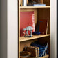 A light wood bookshelf holds books, notebooks, a red canvas, bowls, a blue vase, and the LEGO® Star Wars™ AT-AT™ by Legos - Toyhouse arranged on its shelves.