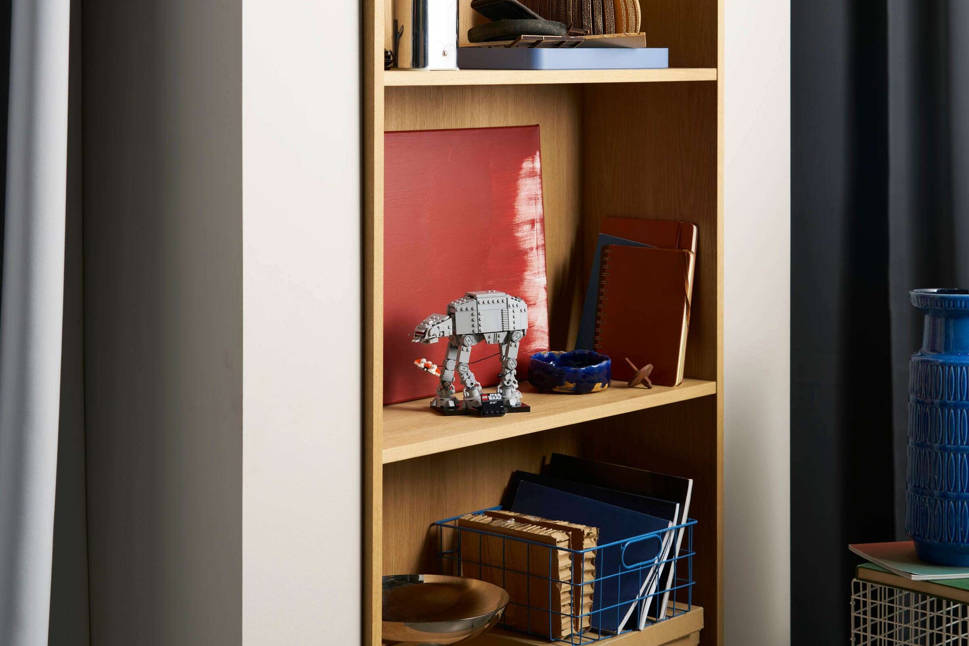 A light wood bookshelf holds books, notebooks, a red canvas, bowls, a blue vase, and the LEGO® Star Wars™ AT-AT™ by Legos - Toyhouse arranged on its shelves.
