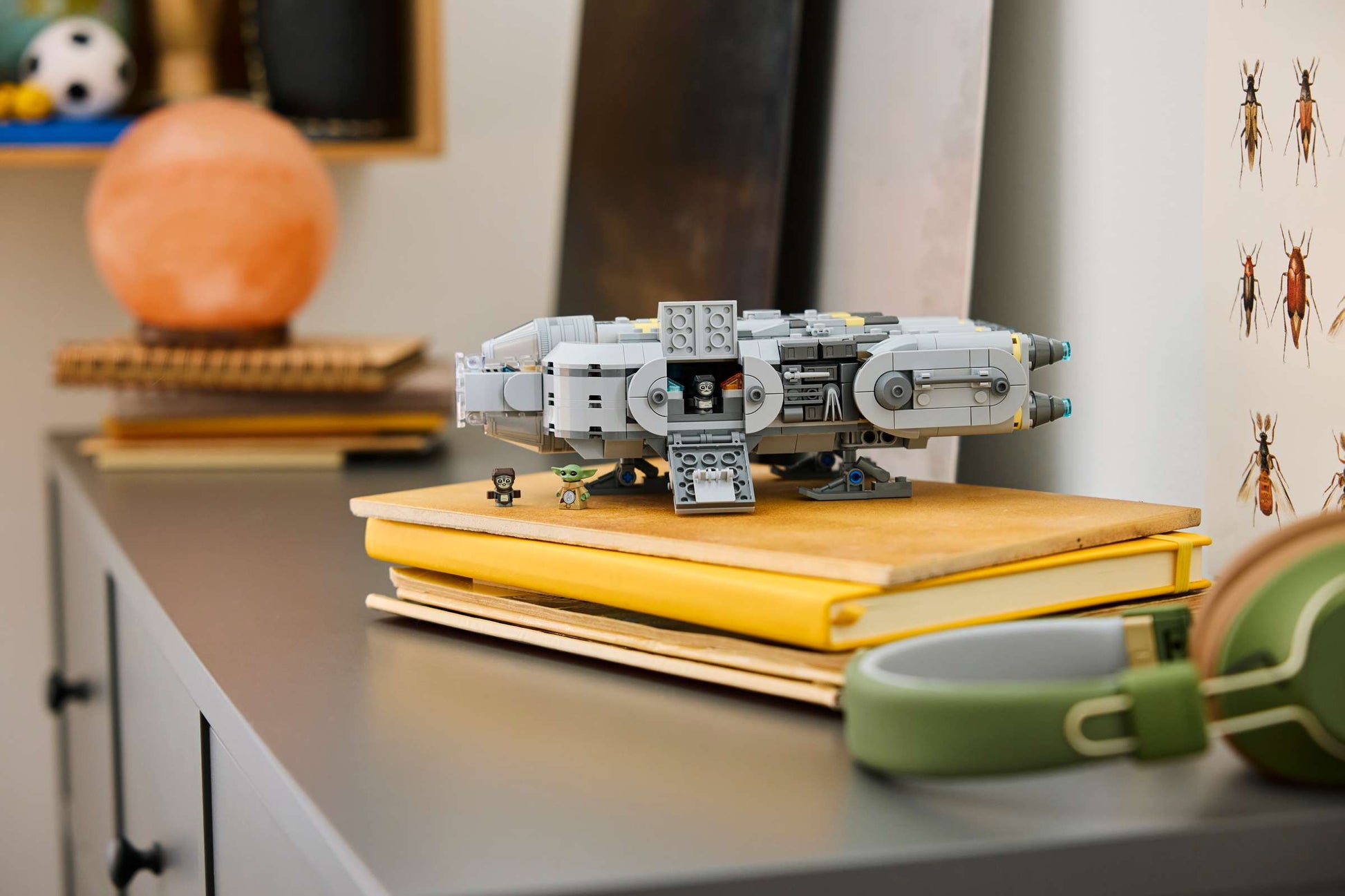 A Legos - Toyhouse LEGO® Star Wars™ Anzellan Starship model rests on yellow books on a gray cabinet, with nearby headphones, a Grogu toy, various objects, and insect illustrations in the background.