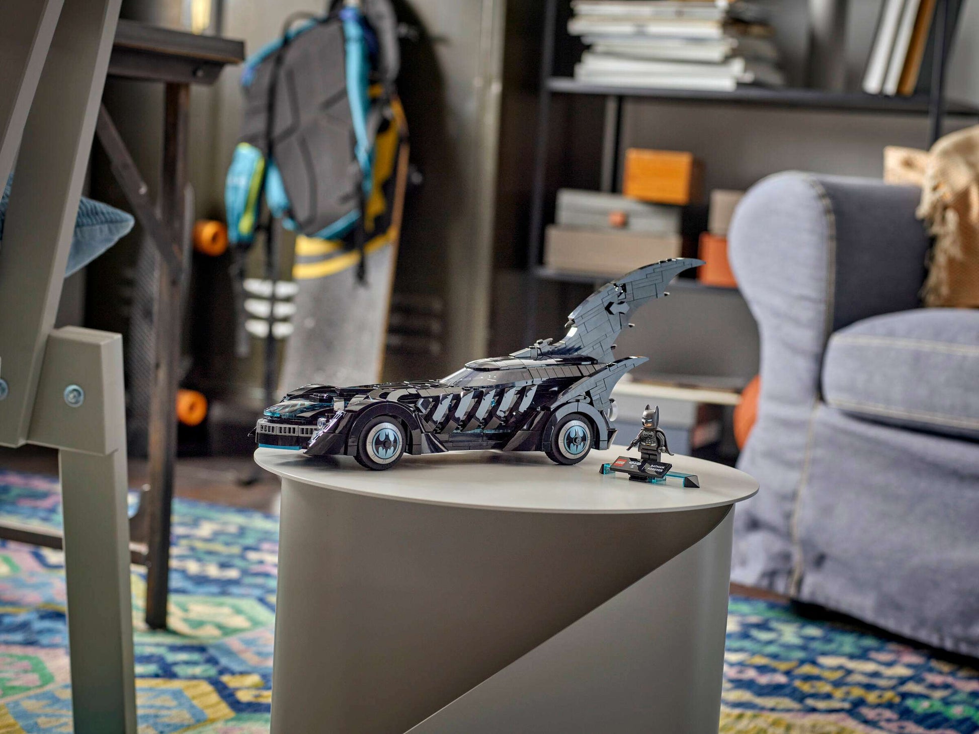 The LEGO® DC Batman Forever™ Batmobile™ and Batman minifigure by Legos - Toyhouse are displayed on a round table in a room with a skateboard, chair, books, and a backpack—ideal for LEGO DC Batman fans.