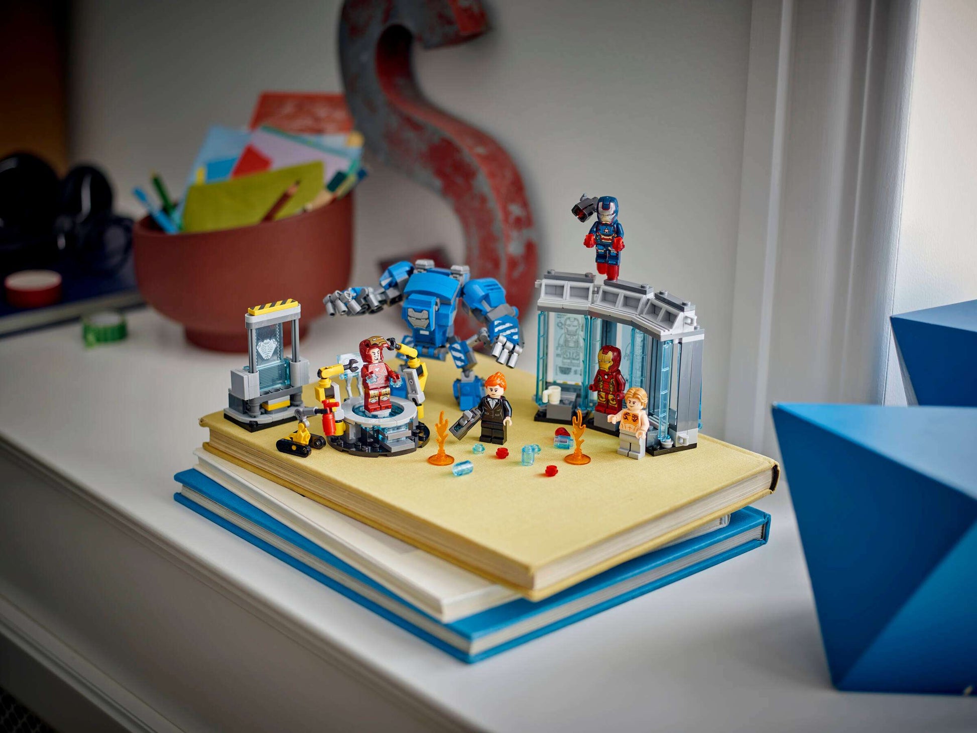 LEGO® Marvel Iron Man's Laboratory: Hall of Armor by Legos - Toyhouse, featuring Iron Man minifigures and accessories, is displayed atop two stacked books on a windowsill with other desk items in the background.