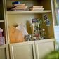 A beige shelving unit holds stacks of paper, a wooden model, a Spider-Man building toy, Legos - Toyhouse sets—including LEGO® Marvel Spider-Man vs. Mysterio: The Daily Bugle—a postcard, and a colorful framed painting partially visible on the right.