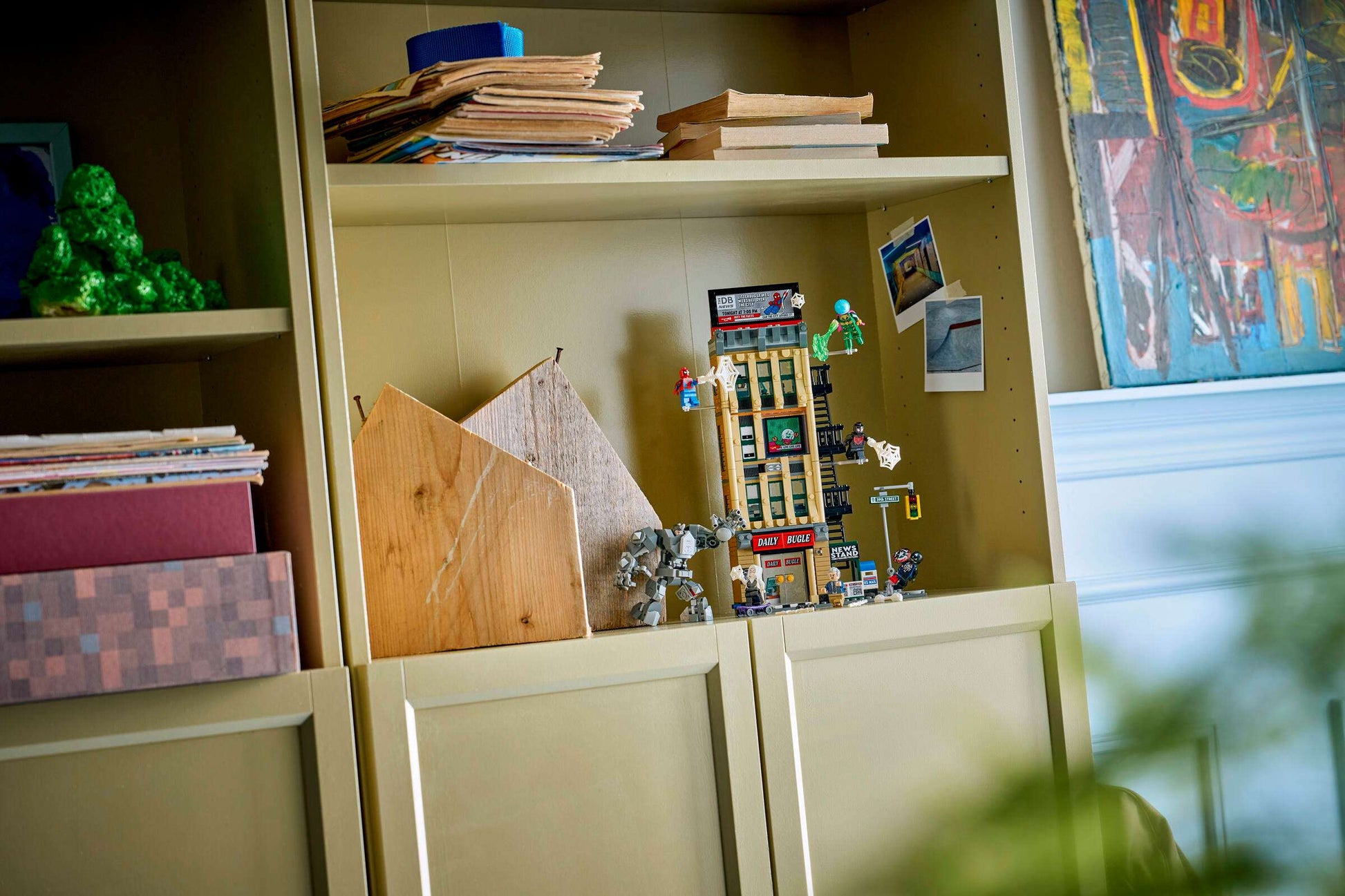 A beige shelving unit holds stacks of paper, a wooden model, a Spider-Man building toy, Legos - Toyhouse sets—including LEGO® Marvel Spider-Man vs. Mysterio: The Daily Bugle—a postcard, and a colorful framed painting partially visible on the right.