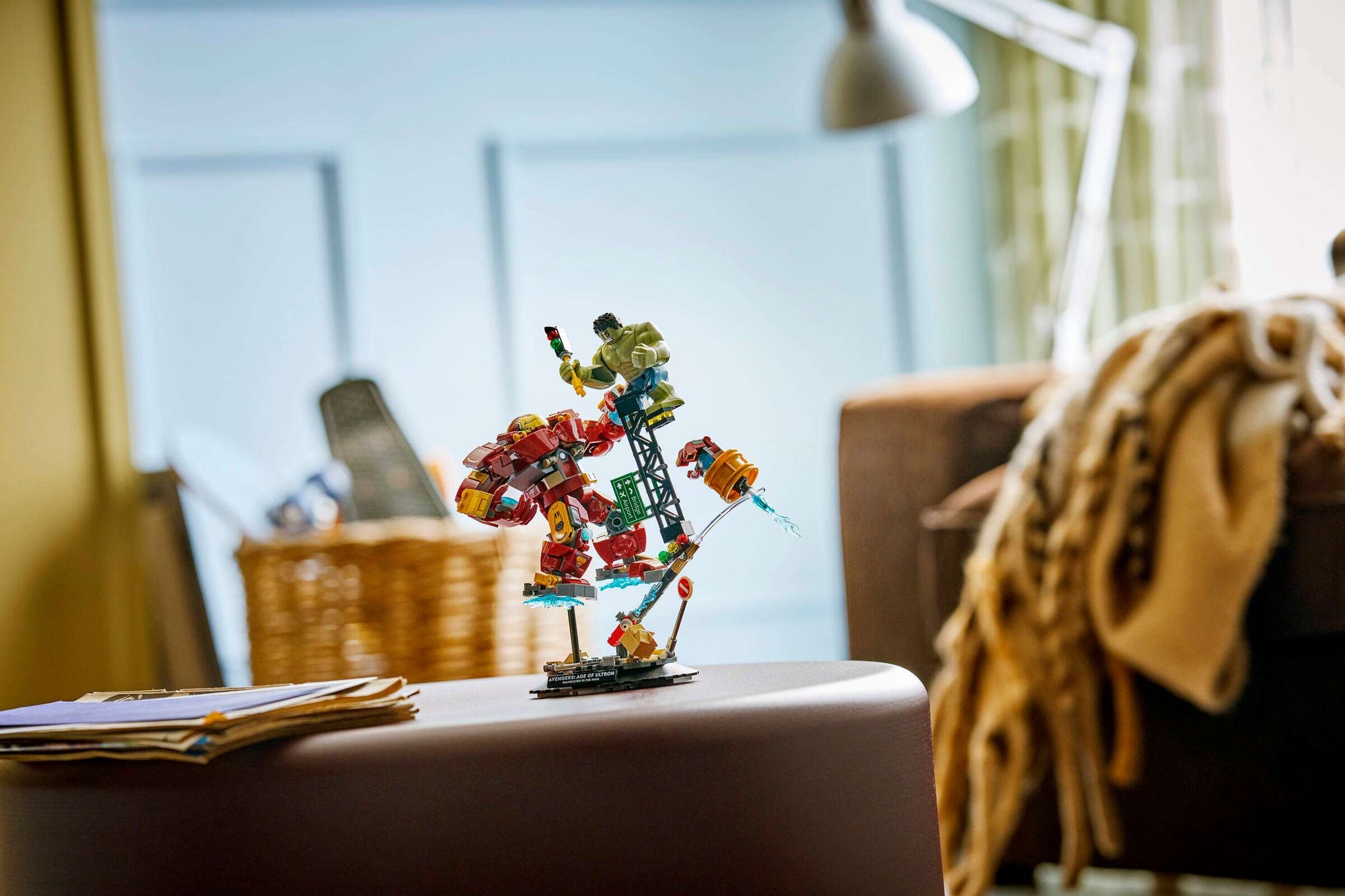 A LEGO® Marvel Epic Battle: Hulkbuster vs. The Hulk display from Legos - Toyhouse is showcased on a round table in a cozy living room, with a blanket, wicker basket, lamp, and papers in the background.