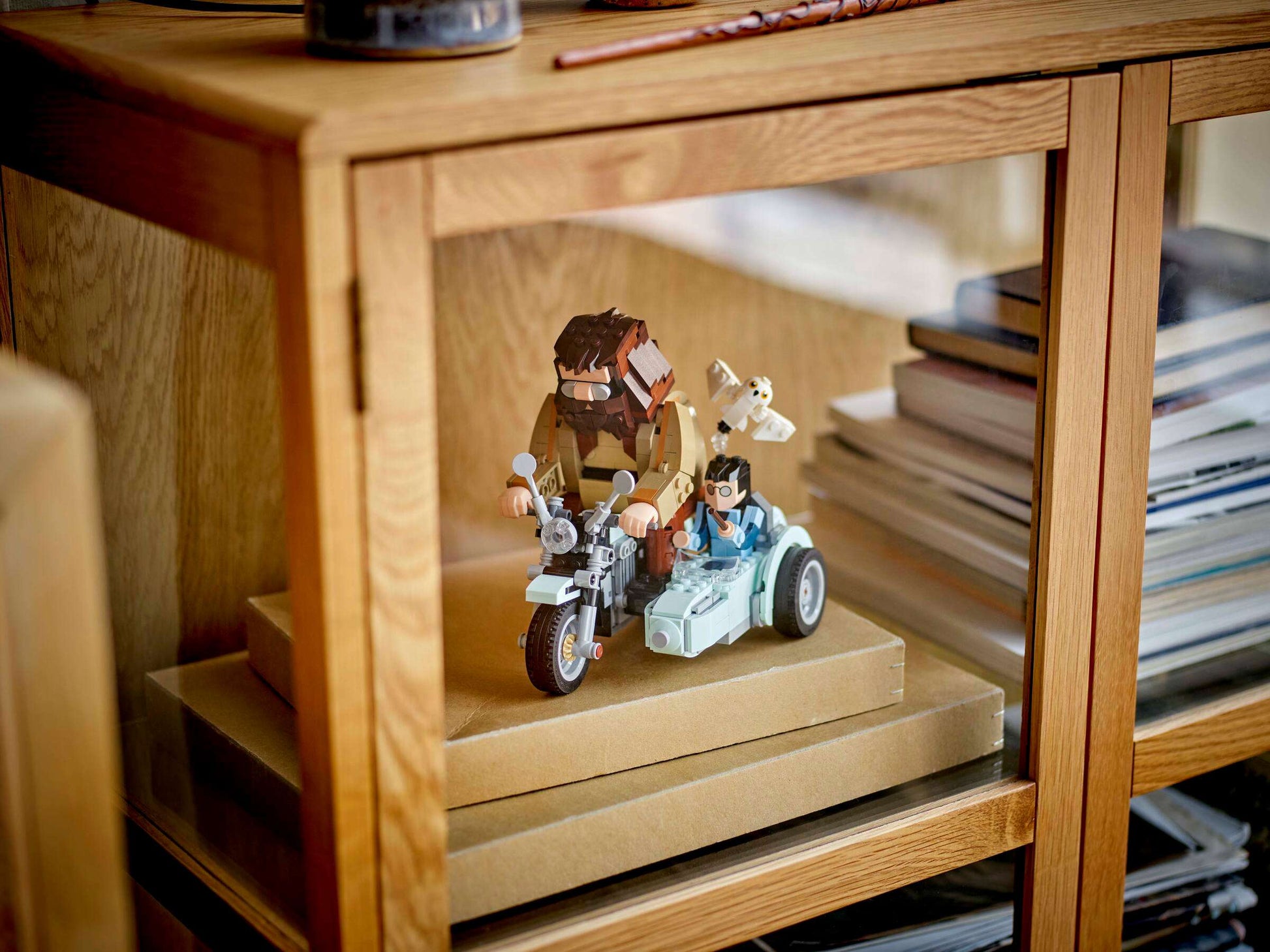 The LEGO® Harry Potter™ Hagrid™ & Harry's Motorcycle Ride set from Legos - Toyhouse is displayed in a wooden glass cabinet, next to stacked books and boxes.