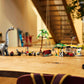 A wooden table displays Legos - Toyhouse LEGO® Jurassic World™ Raptor & Titanosaurus Tracking Mission dinosaur toys and vehicles in a row, with a staircase and indoor plants visible in the background.