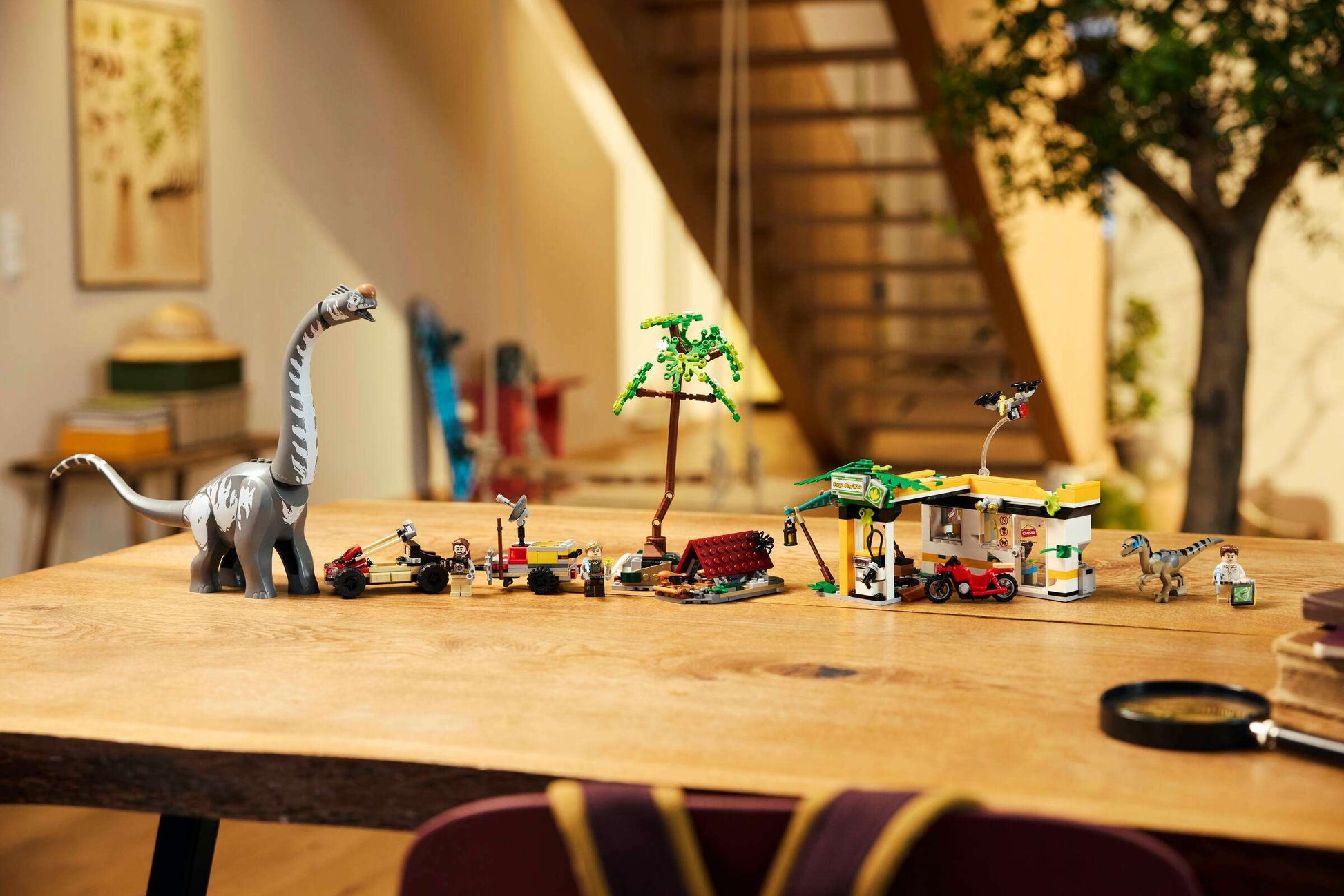 A wooden table displays Legos - Toyhouse LEGO® Jurassic World™ Raptor & Titanosaurus Tracking Mission dinosaur toys and vehicles in a row, with a staircase and indoor plants visible in the background.