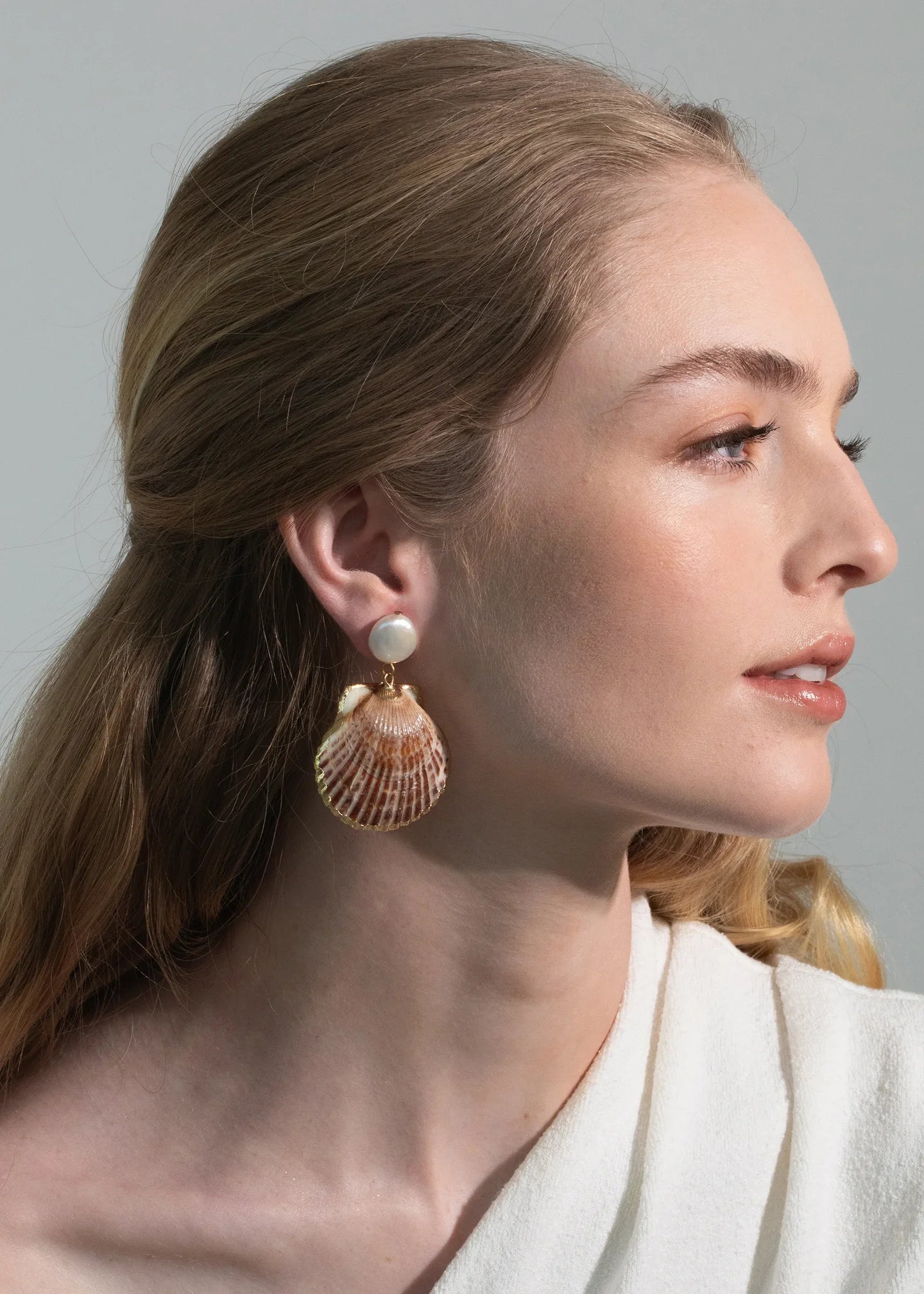 Woman with light brown hair wears Jennifer Behr's Folly Earrings, crafted from natural seashells, and a white top, shown in profile against a neutral background.