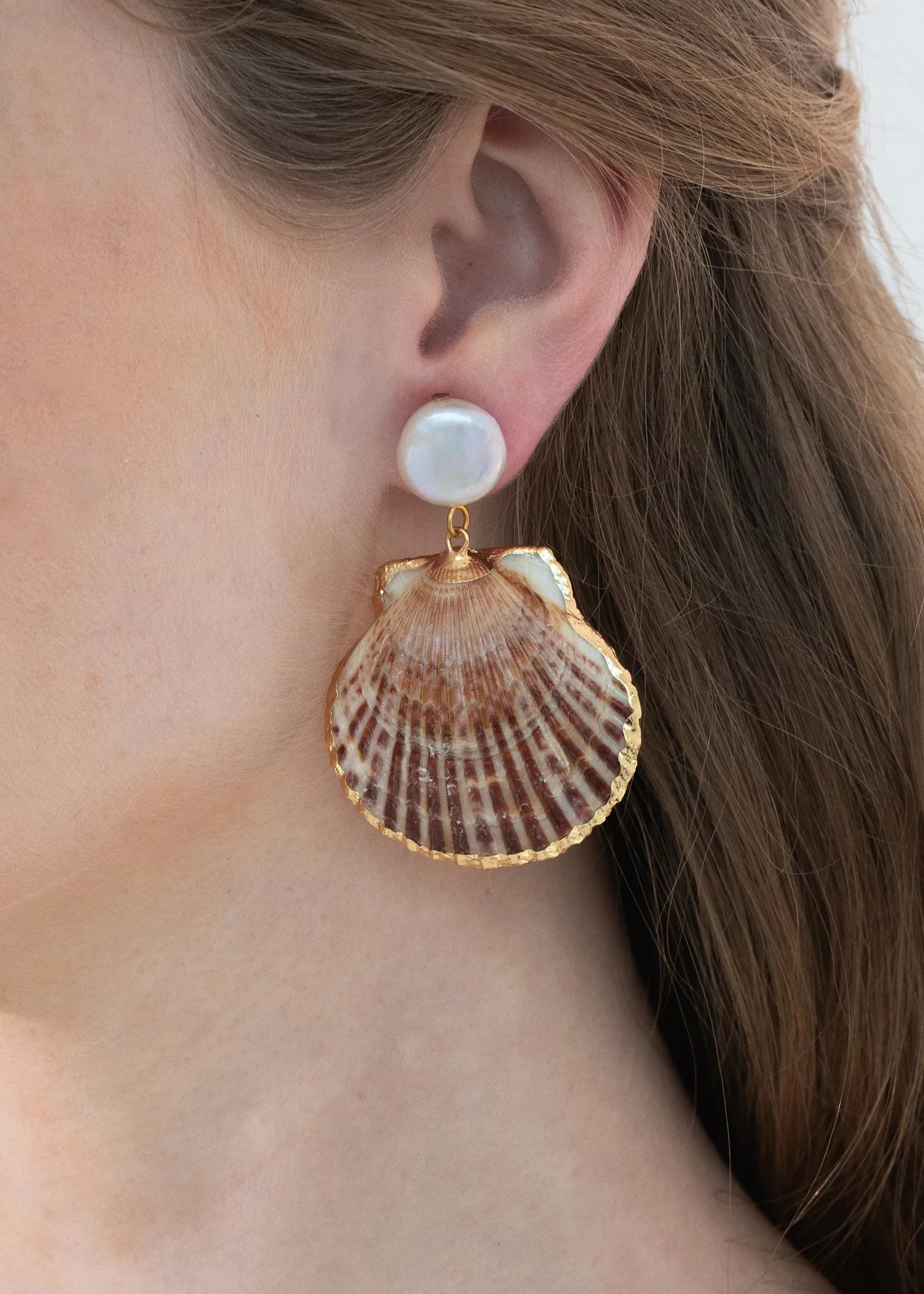 A close-up of a woman's ear wearing Jennifer Behr's Folly Earrings, which feature a white pearl stud with a large natural seashell dangling below.
