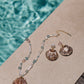 A shell necklace and the Jennifer Behr Folly Earrings, featuring beads and natural seashells, are displayed poolside beside clear blue water.