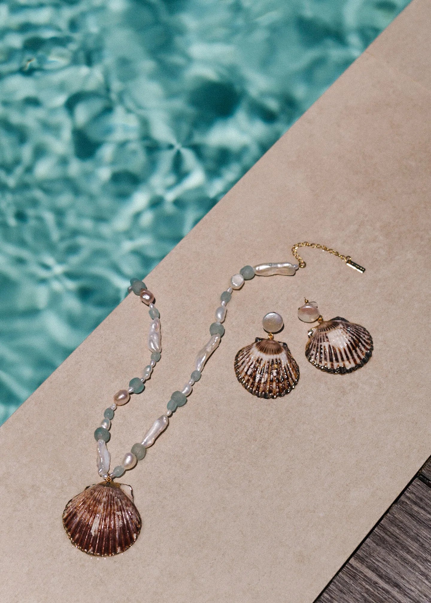 A shell necklace and the Jennifer Behr Folly Earrings, featuring beads and natural seashells, are displayed poolside beside clear blue water.