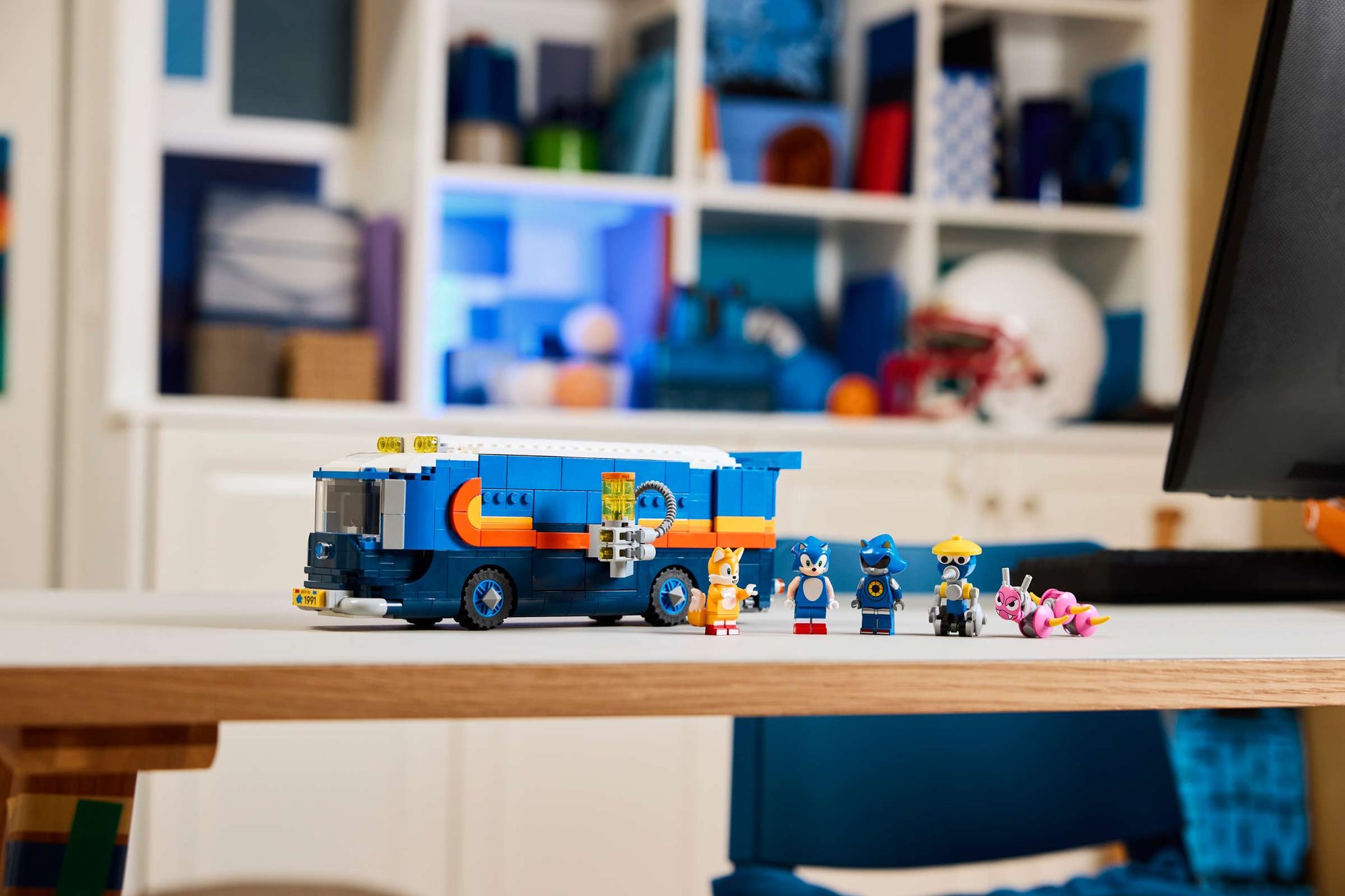 The LEGO® Sonic the Hedgehog™ Team Sonic Command Truck by Legos - Toyhouse features a blue and orange gaming truck and five character figures, shown on a desk in a room with shelves in the background.