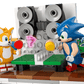 Two LEGO® Sonic the Hedgehog™ Team Sonic Command Truck figures—Tails and Sonic—play table tennis near a speaker wall, with a TV, game controller, and a Sonic toy on a side table. Brand: Legos - Toyhouse.