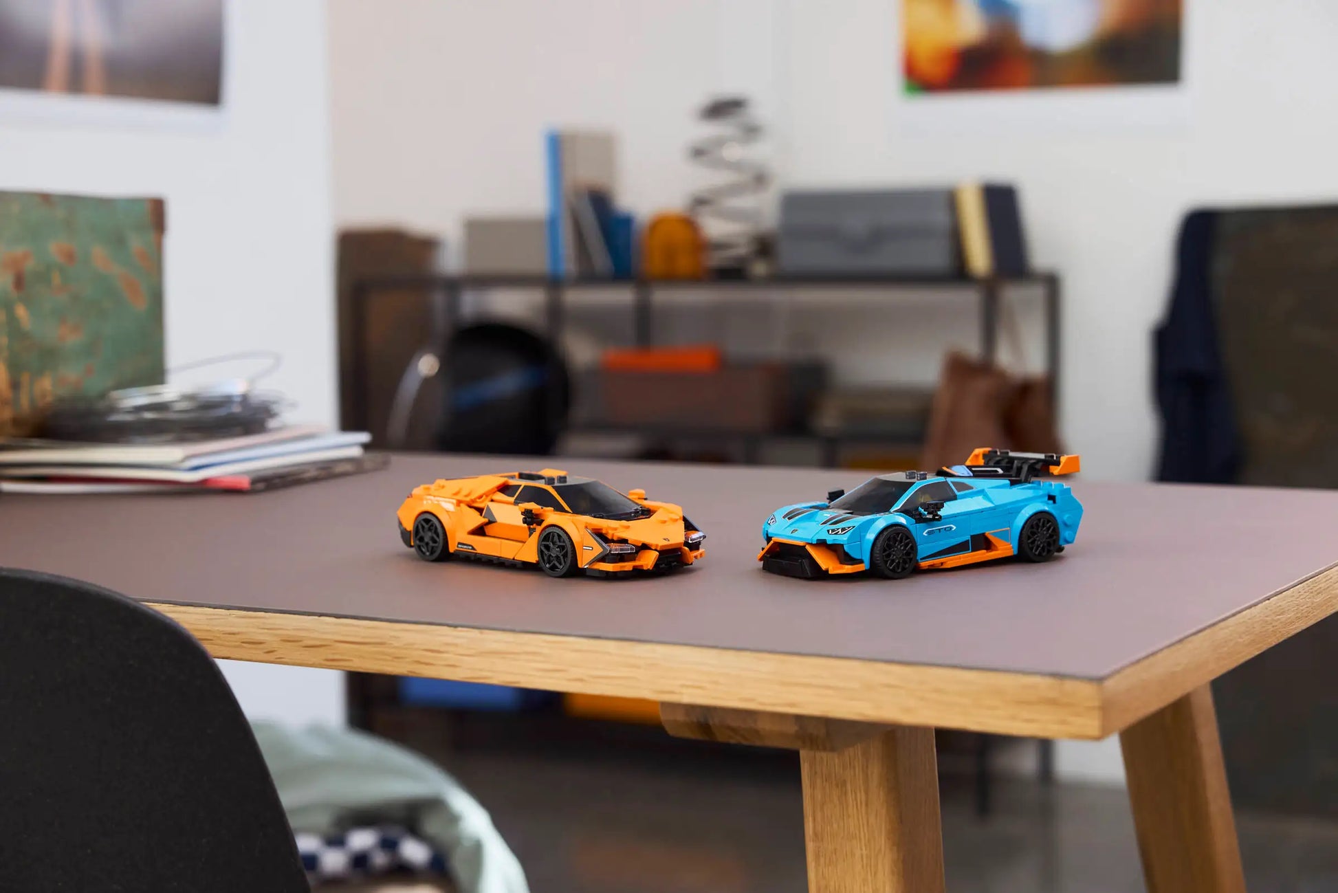 Two LEGO® Speed Champions Lamborghini Revuelto & Huracán STO cars—orange and blue by Legos - Toyhouse—are displayed on a wooden desk in a modern, organized room with shelves and books, ideal for car-building fans.