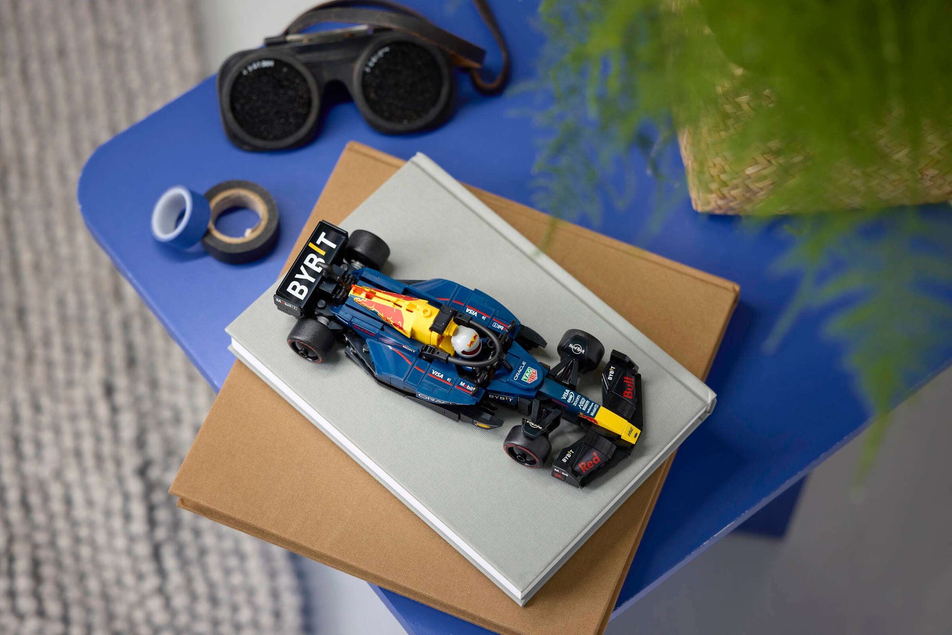 A LEGO® Speed Champions Oracle Red Bull Racing RB20 F1® Race Car by Legos - Toyhouse sits atop two closed books on a blue table, next to goggles, tape, and a green plant.