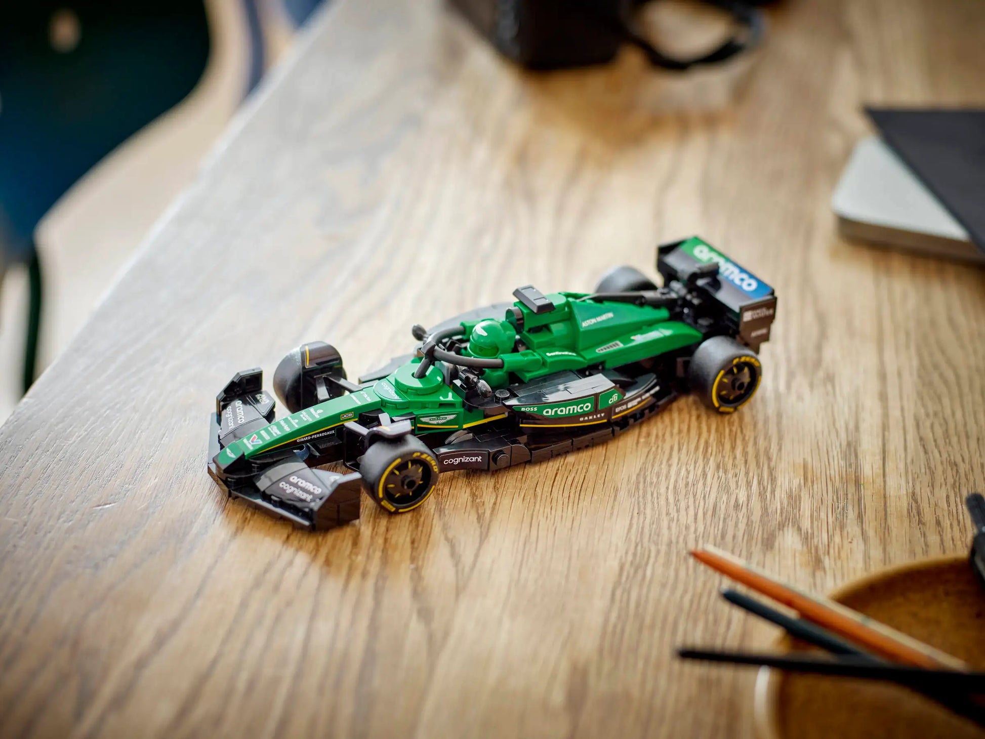 A LEGO® Speed Champions Aston Martin Aramco F1® AMR24 Race Car by Legos - Toyhouse in green and black sits on a wooden table beside pens and a notebook, ideal for Formula 1 fans in 2024.