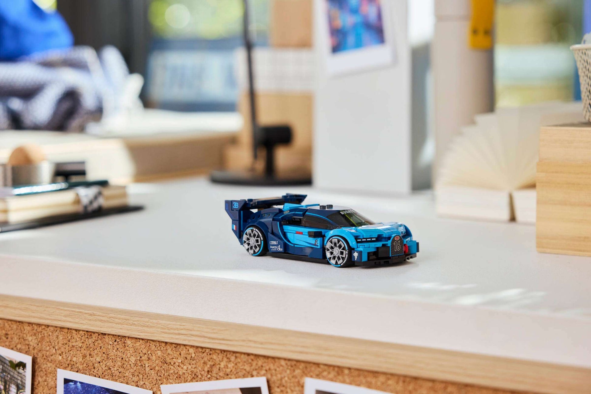 A blue LEGO® Speed Champions Bugatti Vision GT Hyper Sports Car by Legos - Toyhouse sits on a white desk, surrounded by books, photos, and office supplies in the background.