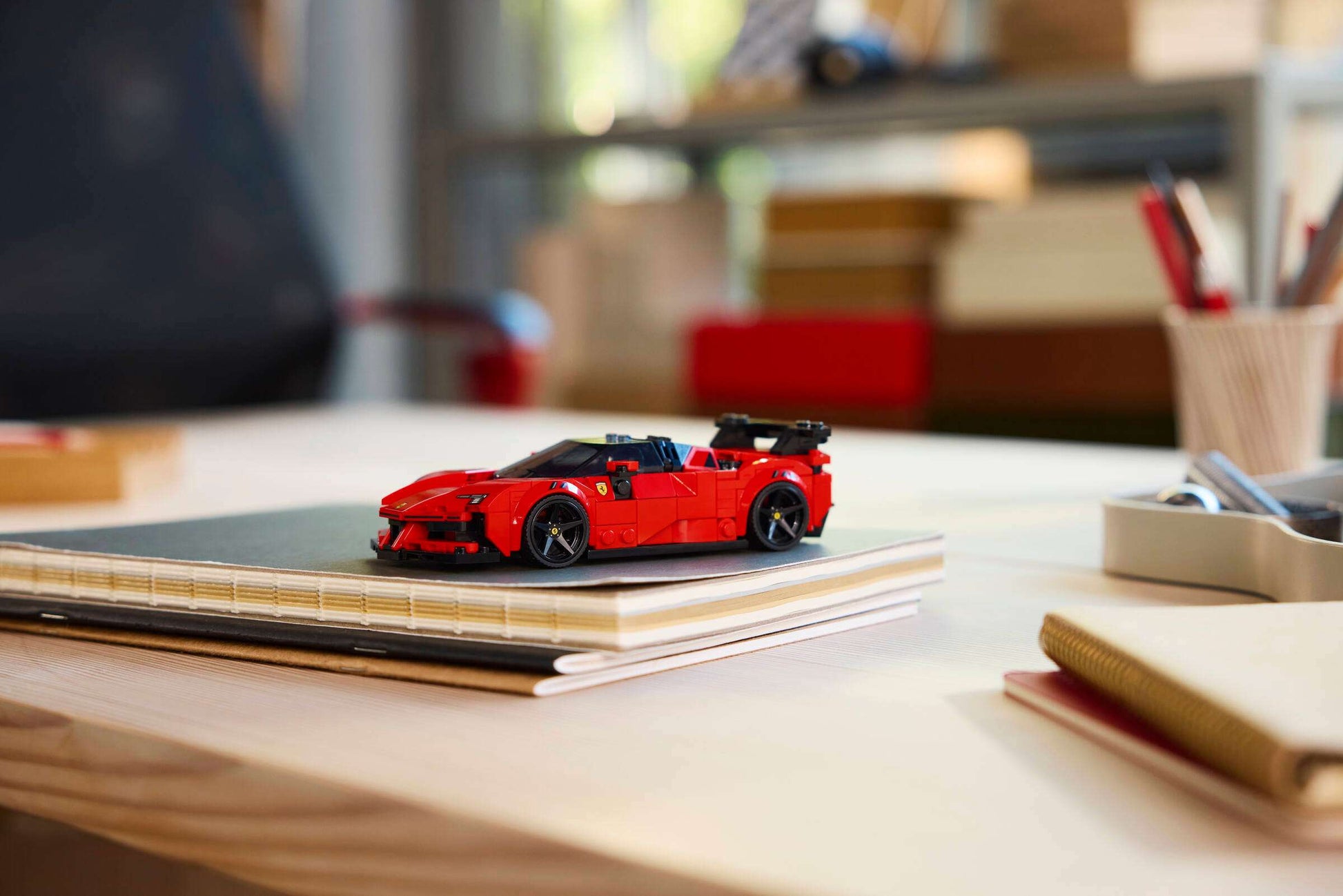 A LEGO® Speed Champions Ferrari SF90 XX Stradale Sports Car by Legos - Toyhouse is displayed on a closed notebook atop a light-colored desk, surrounded by office supplies and blurred shelves in the background.