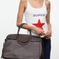 A woman wearing a white tank top with a red star and the word “SUPER” holds the Staud Maude Carryall, an oversized dark brown pebbled leather bag with organizational pockets, against a plain background.