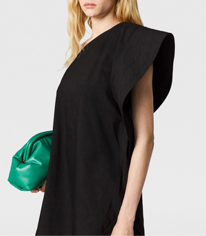 A woman wears the Stella McCartney Washed Cotton Asymmetric Mini Dress in black, holding a green clutch; her head and upper torso are shown against a plain background.