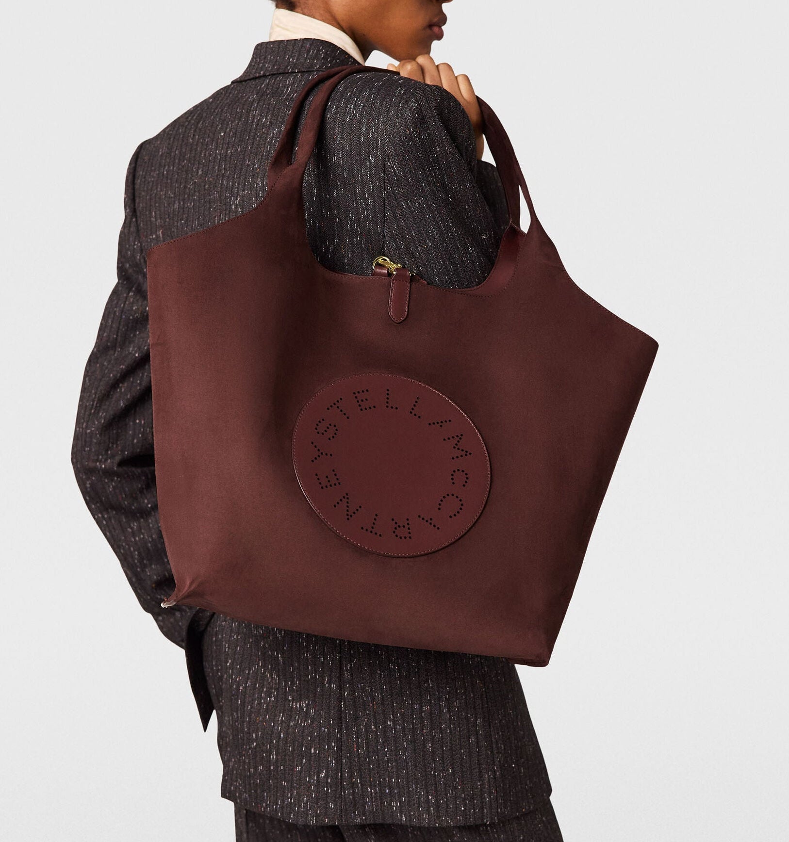 A person in a pinstripe suit holds the Stella McCartney Logo Reversible Suede Tote, featuring perforated “Stella McCartney” lettering.