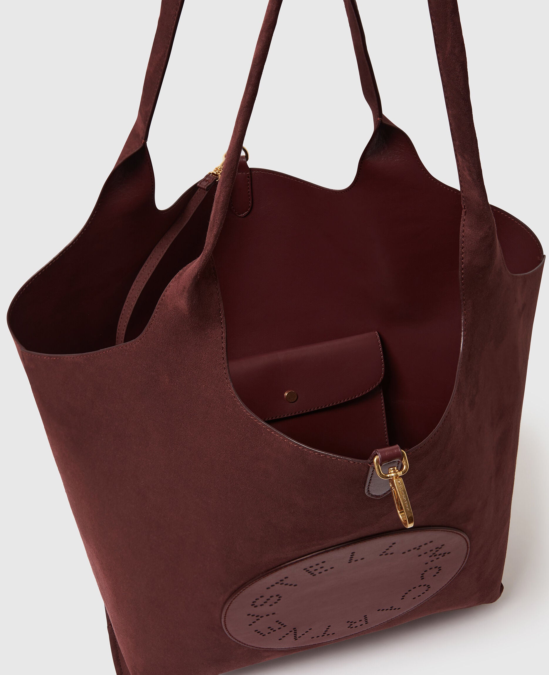 The Stella McCartney Logo Reversible Suede Tote is a spacious burgundy bag featuring a cutout logo, an open top, and an inner pocket with a snap closure.