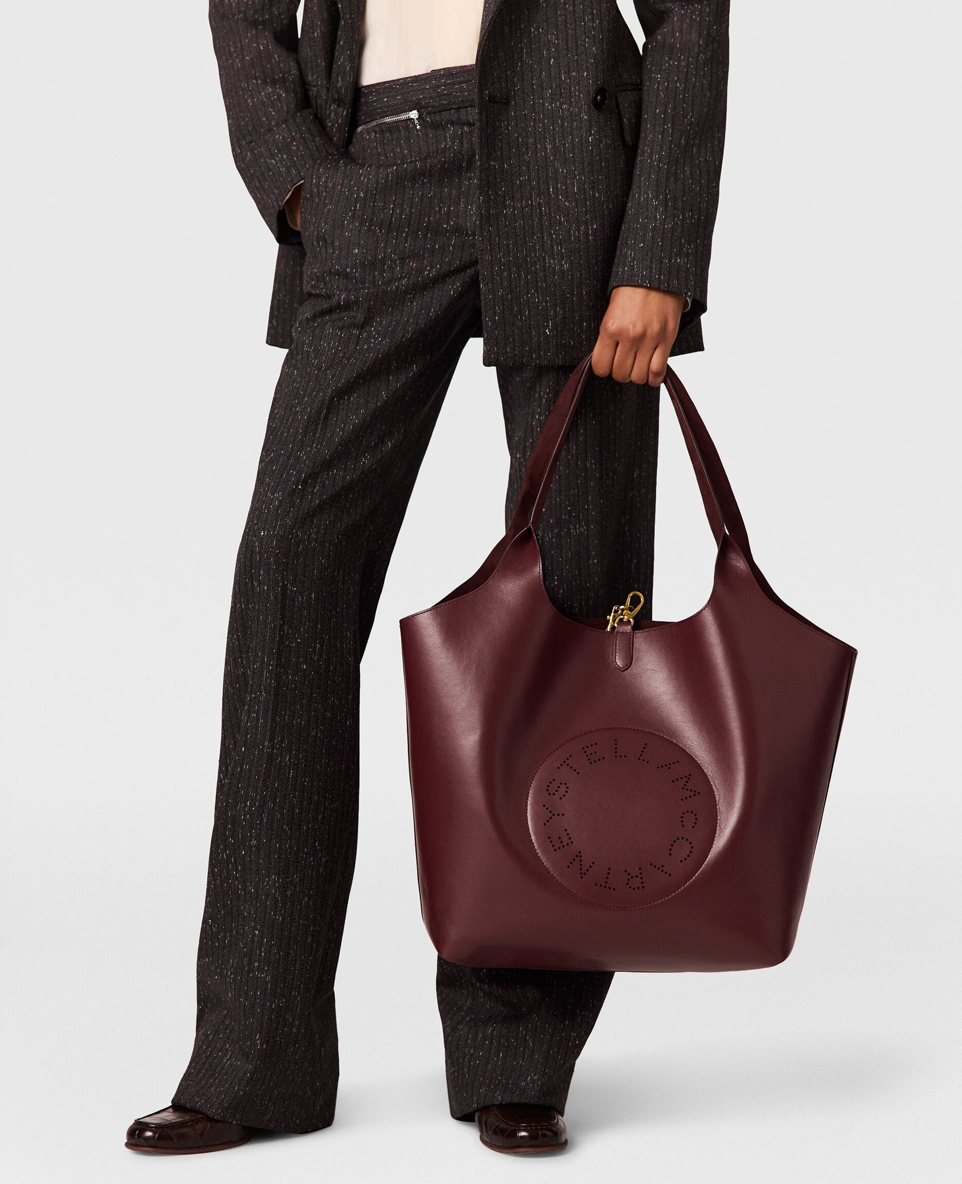 Person in a dark pinstripe suit holding a large burgundy Stella McCartney Logo Reversible Suede Tote.