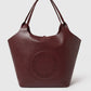 The Stella McCartney Logo Reversible Suede Tote is a large burgundy tote with dual handles and a circular perforated logo on the front, combining versatility and sophisticated design.