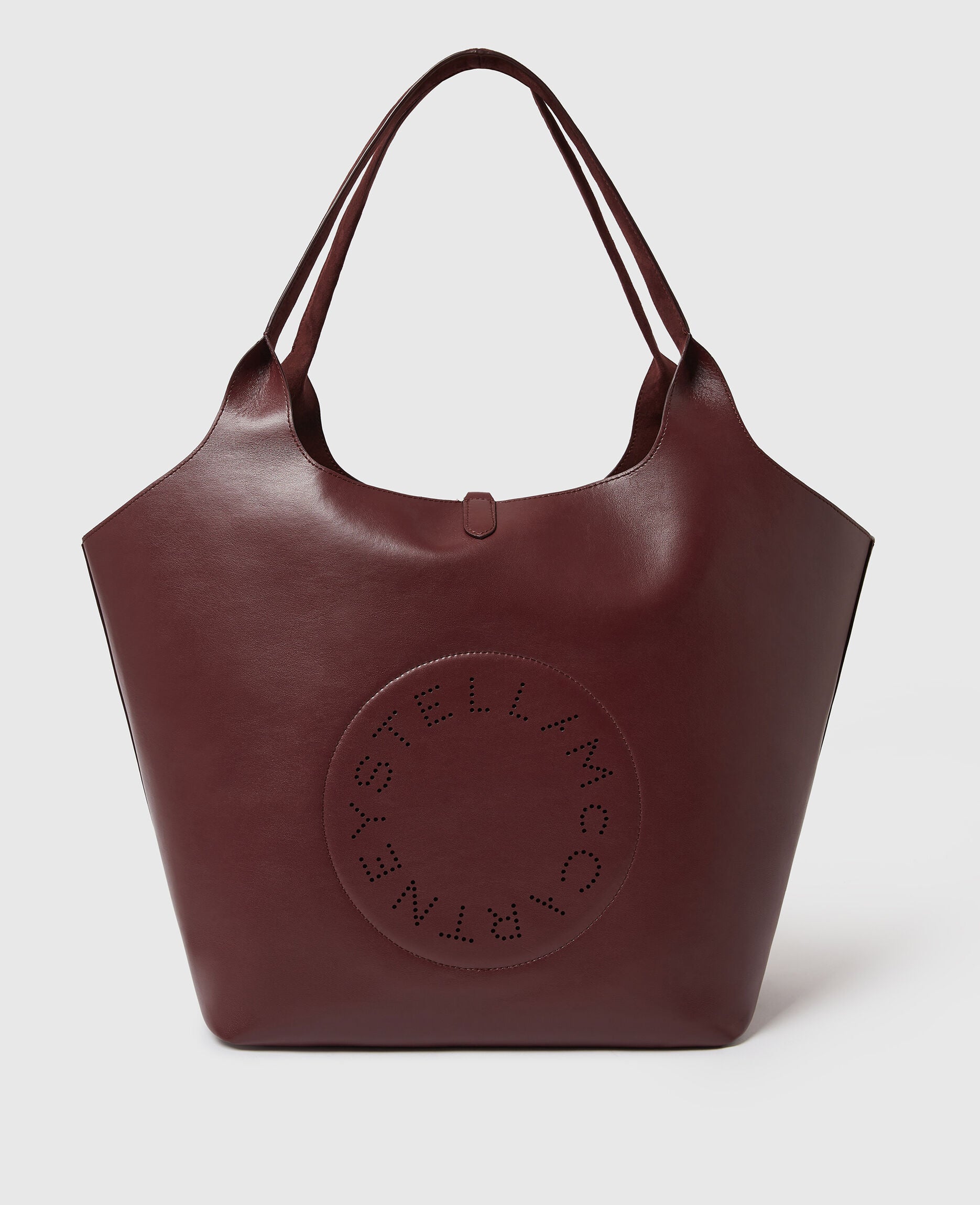 The Stella McCartney Logo Reversible Suede Tote is a large burgundy tote with dual handles and a circular perforated logo on the front, combining versatility and sophisticated design.