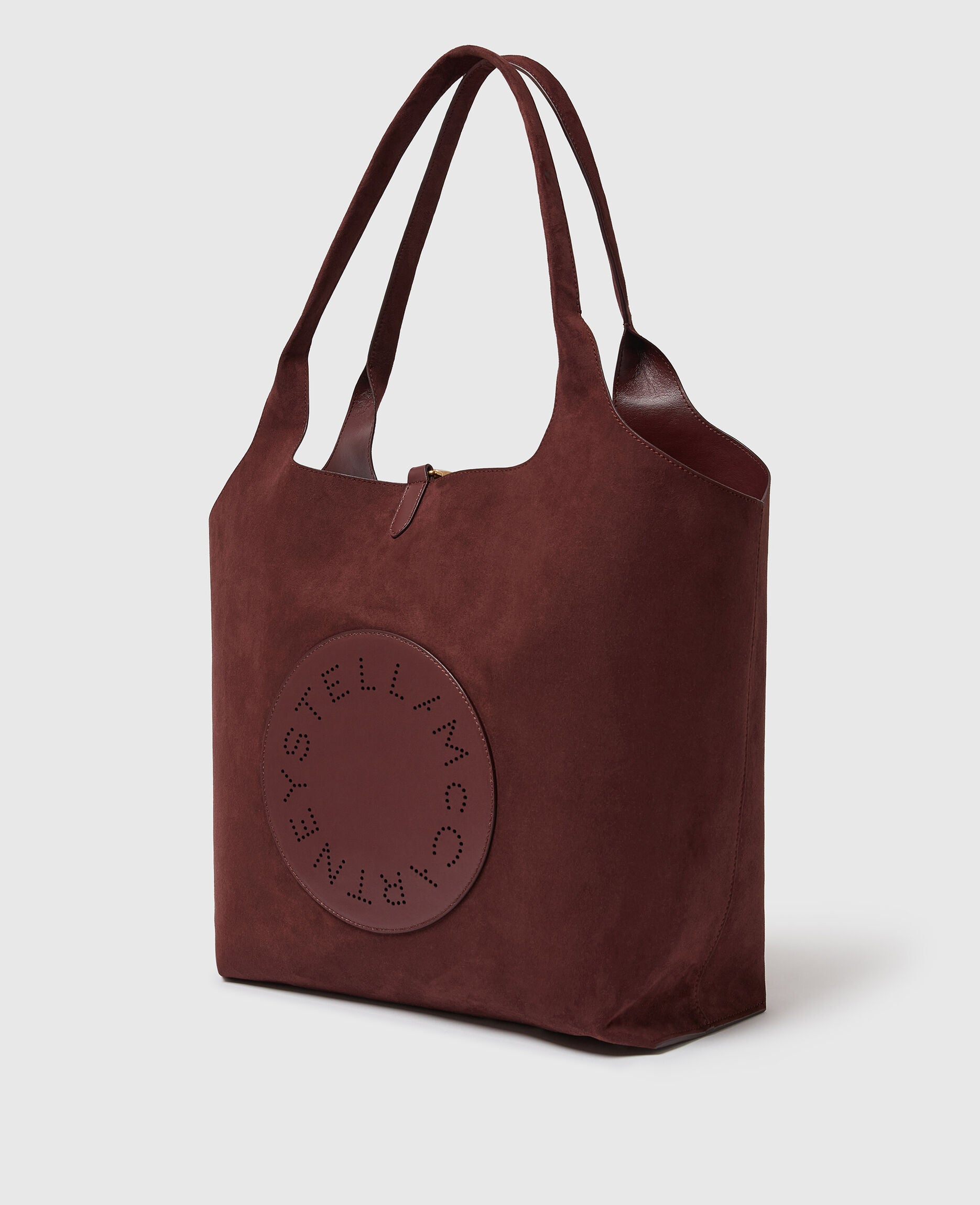 The Logo Reversible Suede Tote by Stella McCartney is crafted in burgundy suede, featuring double handles and a circular front patch with perforated “Stella McCartney” text.