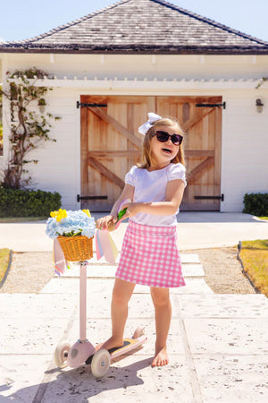 A young girl in sunglasses and a pink checkered skirt stands on a scooter with a flower basket, wearing the Girls' Penny's Play Shirt by The Beaufort Bonnet Company, in front of a white garage with wooden doors.