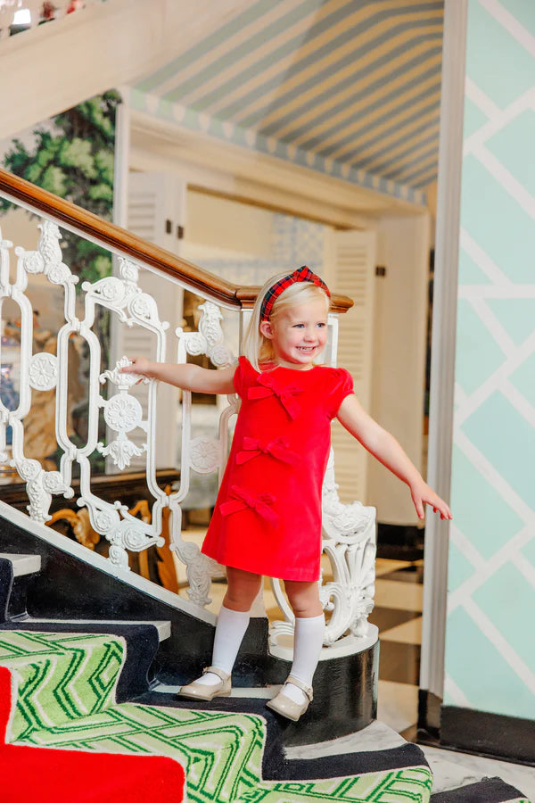 A young girl in a Girls' Velveteen Betts Bow Dress by The Beaufort Bonnet Company stands smiling on a staircase with patterned carpet and an ornate white railing.