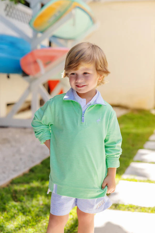 A young boy in The Beaufort Bonnet Company’s Baby Boys' and Boys' Prepletic™ Hayword Half Zip stands outside smiling, with colorful kayaks stacked in the background.