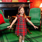 A young girl in a red Girls' Darcy Dress by The Beaufort Bonnet Company stands with arms outstretched on a green plaid carpet, surrounded by green chairs and a cozy fireplace.